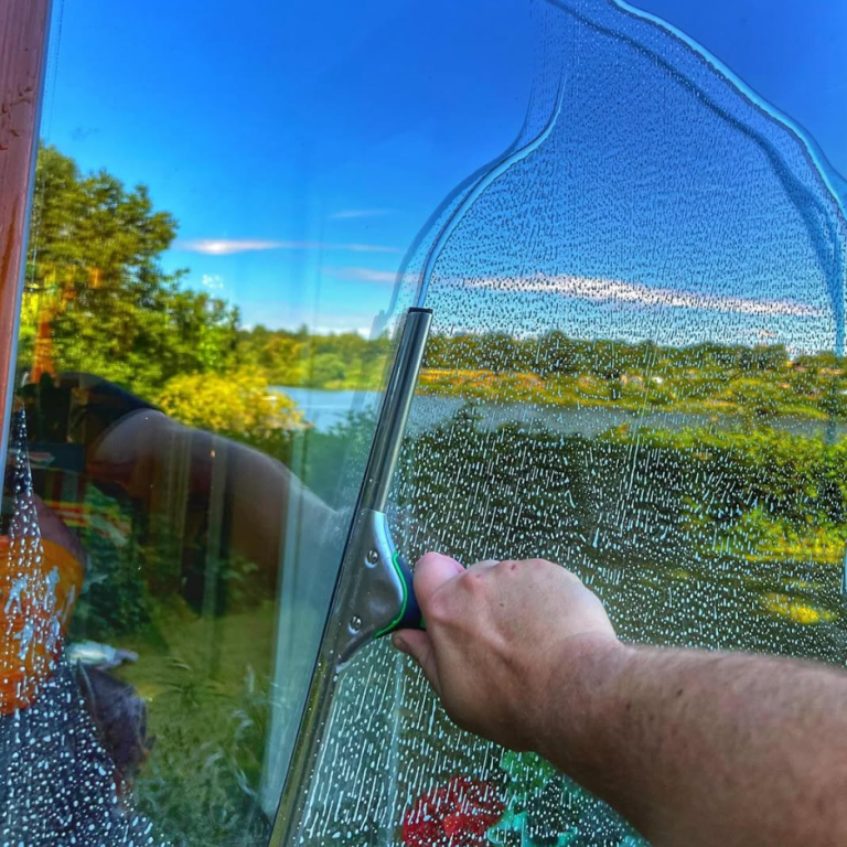 Fensterreinigung Eine Hand reinigt ein beschlagenes Fenster mit Blick auf einen See und die umliegende Natur.